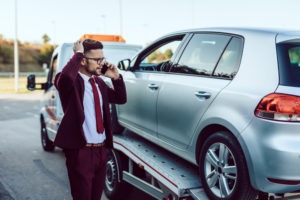 An upset businessman on his phone and standing next to his car that’s being towed.